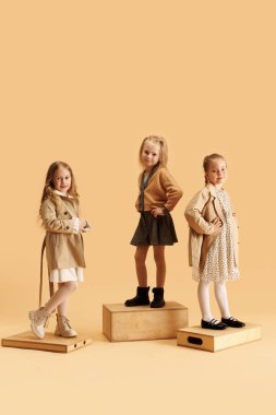 Üç küçük kız, bej renkli stüdyo arka planında kameraya bakan çekici, sıcak bir sonbahar kıyafeti giymiş. Moda ve tarz kavramı, güzellik, çocukluk, Kara Cuma, Siber Pazartesi.