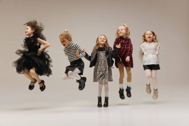 Neşeli küçük kızların dinamik portresi. Gri stüdyo arka planına karşı şık kıyafetler içinde mutluluktan zıplıyorlar. Çocukluk kavramı, çocuk modası, güzellik, tarz, okul alışverişi. Kara Cuma