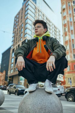 Genç adam hareketli şehir manzarasına karşı poz veriyor, neon büyüklüğünde elbisesi modern sokak modasının cesur ifadesi. Sokak modası ve şehir tarzı kavramı, modern yaşam tarzı, Z jenerasyonu, kendini ifade etme.