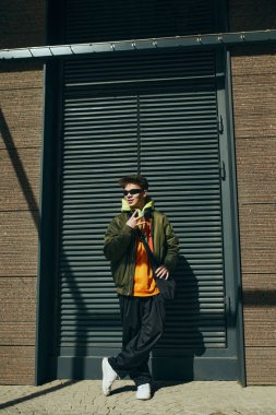 Modayı belirleyen genç ve çekici adamın modern portresi, konfor ve kentsel modanın bina yakınında duran birleşimini sergiliyor. Sokak modası ve şehir tarzı kavramı, modern yaşam tarzı, Z jenerasyonu.