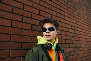 Canlı kapüşonlu ve kulaklık takan şık bir adamın tasviri. Duvara karşı açık havada poz veriyor. Sokak modası, şehir tarzı, modern yaşam tarzı, Z jenerasyonu, kendini ifade etme.