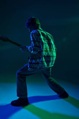 Retro kıyafetli genç artistik gitarist. Gradyan stüdyo arka planına karşı yeşil mavi sahne ışığı altında gösteri yapıyor. Müzik ve sanat kavramı, hobi, konserler ve festivaller, modern kültür. Ad