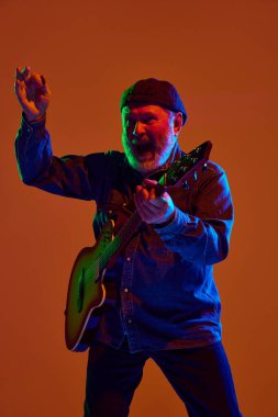 Sakallı yaşlı müzisyen turuncu stüdyo arka planına karşı renkli neon ışıkta elektro gitar çalıyor. Müzik ve sanat kavramı, hobi, konserler ve festivaller, modern kültürün son sınıfları. Ad