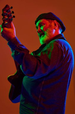 Gayri resmi giyinen yaşlı gitarist turuncu stüdyo arka planında renkli neon ışıkta performans sergiliyor. Müzik ve sanat kavramı, hobi, konserler ve festivaller, modern kültürün son sınıfları. Ad