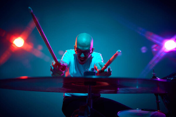 Emotional bald drummer in casual outfit with drumsticks in pink-purple stage lighting against gradient studio background. Concept of music and art, hobby, concerts and festivals, modern culture. Ad