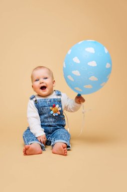 Kot pantolon giymiş keyifli bir bebek bulutlarla süslenmiş mavi bir balon tutuyor ve sıcak bej arka planda gülüyor. Çocukluk kavramı, annelik, yaşam, doğum. İlan için alanı kopyala