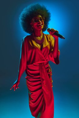 Canlı retro-moda elbisesi ve güneş gözlüklü neşeli Afrikalı-Amerikalı şarkıcı, arka planda neon ışığı altında performans sergiliyor. Sanat, müzik, hobi, klasik müzik ve modern yaşam tarzı kavramı.