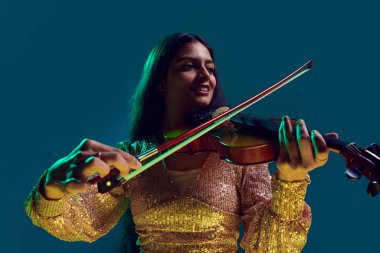Smiling Indian female violinist in shimmering crop top with serene expression in neon light against gradient background. Concept of art, music, hobby, classical music and modern lifestyle. Ad clipart