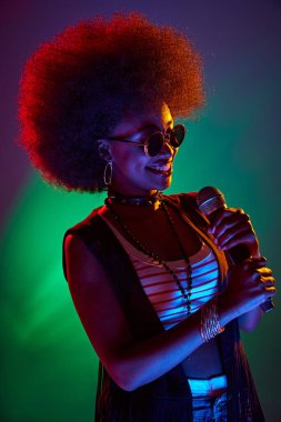 Büyük afro ve retro elbiseli bayan şarkıcının portresi, mikrofon tutuyor, neon ışıkta, arka planda eğilim gösteriyor. Sanat, müzik, hobi, klasik müzik ve modern yaşam tarzı kavramı. Ad