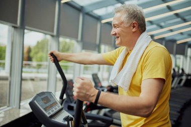 Canlı spor kıyafetleri içinde, omuzlarında havluyla gülümseyen son sınıf öğrencisi egzersiz makinesi modern spor salonunda eğitim görüyor. Spor anlayışı, modern hayatın aktif yaşlıları, sağlıklı yaşam tarzı. Ad