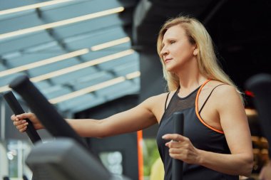 Antrenman kıyafeti giymiş, eliptik antrenör kullanan olgun bir kadın modern spor salonundaki fitness rutinine odaklanmış. Spor anlayışı, modern hayatın aktif yaşlıları, sağlıklı yaşam tarzı. Ad