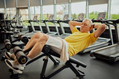 Emekliler için modern fitness merkezinde abdominal egzersizler yapan enerjik spor kıyafetleri içindeki senyor adamı eğitmeye odaklanmış. Spor anlayışı, modern hayatın aktif yaşlıları, sağlıklı yaşam tarzı. Ad