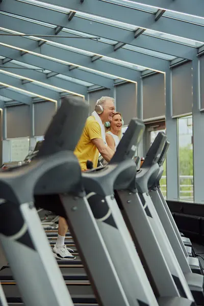 Laughing senior couple enjoys fun workout on treadmills in modern fitness center. Cardio training to fat burning. Concept of sport, active seniors in modern life, healthy lifestyle. Ad