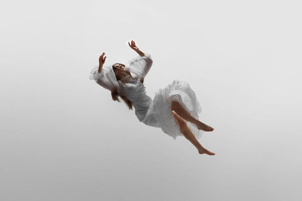 Levitating dream. Dynamic photo of young woman in white elegant gown, rising levitating in mid-air against white studio background. Concept of beauty, feminine elegance and purity, dream and reality.
