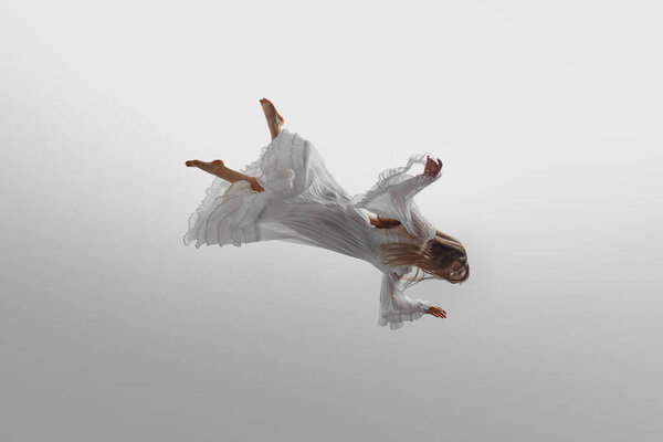 Woman dressed in vintage white gown suspend in mid-air, her long blonde hair cascading around her against white studio background. Concept of beauty, feminine elegance and purity, dream and reality.