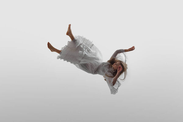 Elegantly dressed in white vintage gown, woman floats, falling down in mid-air in delicate pose, against white studio background. Concept of beauty, feminine elegance and purity, dream and reality.