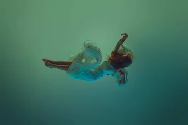 Young woman dressed in white gown elegantly dancing or descent through water against serene aqua background. Concept of underwater fantasy, freedom and weightlessness, mystery and depth. Ad