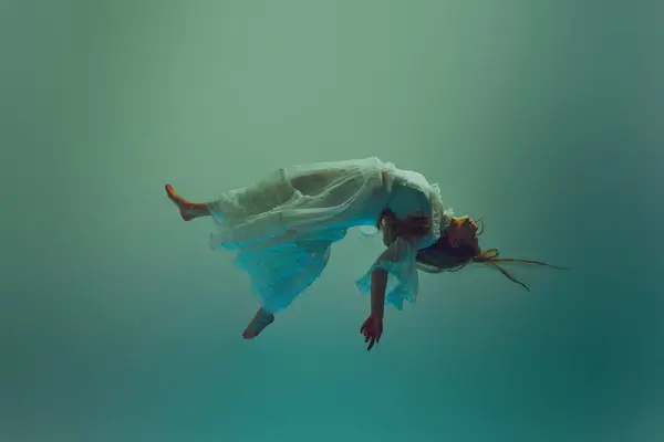 Young woman in flowing vintage white dress in motion, she moved softly under invisible stream of water. Model levitating against aqua background. Concept of beauty, elegance, mystery and depth. Ad