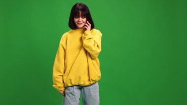 Moda kıyafeti giymiş genç bir kadın telefonda konuşuyor ve canlı yeşil stüdyo geçmişine gülümsüyor. İnsan duyguları kavramı, moda, güzellik, tarz, mesafe iletişimi.