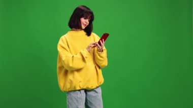 Moda kıyafeti giymiş genç bir kadın telefonla konuşuyor ve canlı yeşil stüdyo geçmişine gülümsüyor. İnsan duyguları kavramı, moda, güzellik, tarz, mesafe iletişimi