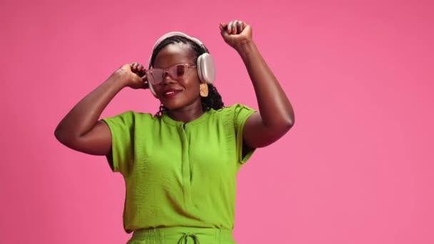リラックスしたアフリカ系アメリカ人女性がヘッドフォンで音楽を聴き パステル ピンクのスタジオのバックグラウンドで歌います 人間の感情 ファッション 美しさ トレンド パーティー モダンなライフスタイルの概念 — ストック動画