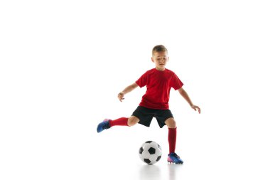 Spor üniformalı küçük bir çocuğun dinamik çekimi futbol oynuyor. Beyazların arka planında pas atma antrenmanı yapıyor. Profesyonel spor, şampiyonluk, gençlik ligi, hobi. Ad