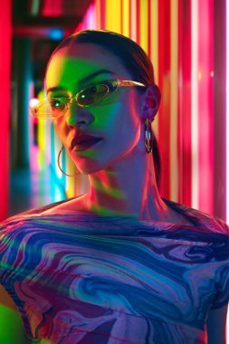 Modern güneş gözlükleriyle süslenmiş, neon çizgilerin canlı arkaplanına karşı karışık parlak bir ışık altında poz veren modacı kadın. Gençlik kavramı, güzellik, moda ve tarz, modern yaşam tarzı. Ad