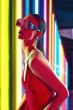 Çarpıcı kırmızı elbiseli ve fütürist güneş gözlüklü kadın portresi onun cesur ve kendinden emin duruşunu vurgulayan yoğun renkleri yansıtıyor. Gençlik kavramı, güzellik, moda ve tarz, modern yaşam tarzı. Ad