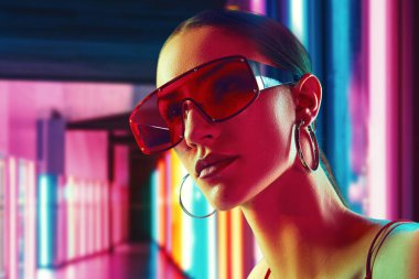 Modayı şehir cazibesiyle harmanlamak. Parlak, modern aksesuarlarıyla neon ışıklı odada poz veren genç ve güzel bir kadının portresi. Gençlik kavramı, güzellik, moda, tarz, modern yaşam tarzı
