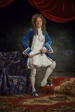 Ortaçağ insanı, eski moda kıyafetler giyen yaşlı adam klasik stüdyo geçmişine karşı kılıçla poz veriyor. Çağların karşılaştırmaları, modernlik ve tarihin birleşimi, moda ve tarz kavramı