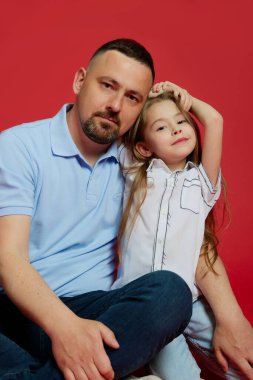 Babasının portresi, sevgili küçük kızıyla birlikte canlı kırmızı arka planda poz vererek günlük giysiler giymişti. Babalar Günü kavramı. Çocuk günü, aile günü, ebeveynlik. Ad