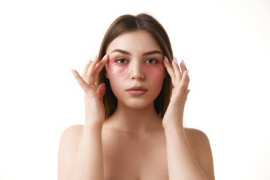 Portrait of young brunette woman with natural makeup look and bare shoulders applying hydrogel eye patches against white studio background. Concept of natural beauty and appearance, facial care. clipart