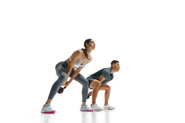 Genç adam ve kadının dinamik fotoğrafı beyaz stüdyo arka planına karşı çömelmiş vaziyette kettlebells ile egzersiz yaparken görülüyor. Spor, sağlıklı yaşam tarzı, takım çalışması, motivasyon. Ad
