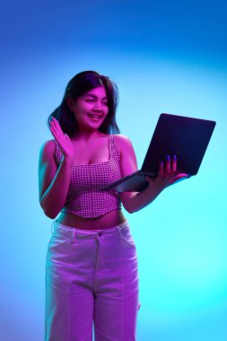 Neşeli genç Hintli kız, öğrenci pembe neon ışıkta mavi arka planda dizüstü bilgisayara bakarken el sallıyor. İnsan duyguları kavramı, online çalışma ve çalışma, gençlik. Ad