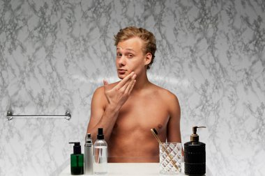 Genç üstsüz adam banyoda dikiliyor ve mermer duvara karşı derin düşünceli bir ifadeyle aynaya bakıyor. Erkek sağlığı, güzellik ve kendine bakım, gençlik, vücut ve cilt bakımı kavramı.