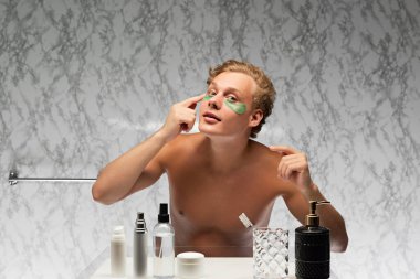 Young shirtless man applying eye patches to moisturize preorbital zone against marbled wall in bathroom. Remove swelling. Concept of male health, beauty and self care, youth, body and skin care. clipart