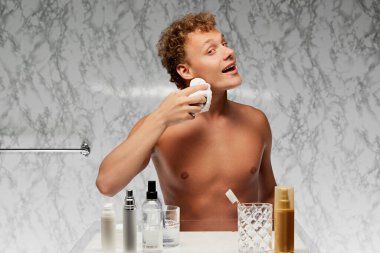 Genç üstsüz bir adam banyoda mermer duvarda tıraş olduktan sonra yüzünü temizlemek için yüz süngeri ya da bez kullanıyor. Erkek sağlığı, güzellik ve kendine bakım, gençlik, vücut ve cilt bakımı kavramı.