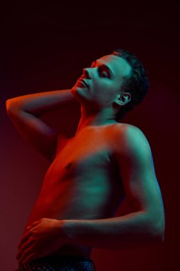 Bir anlık sakinlik. Genç çekici bir adam, arka planda neon ışıkta elini boynunun arkasında tutarak üstsüz poz veriyor. Erkek sağlığı, güzellik ve kendine bakım, gençlik, vücut ve cilt bakımı kavramı.