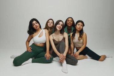 Vücut tiplerini ve etnik kökenlerini temsil eden kadınlar, beyaz arka plana karşı şık atletik kıyafetler içinde birlikte poz veriyorlar. Güzellik ve moda kavramı, destek, birlik, sağlık, vücut pozitifliği..