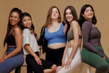 Farklı etnik kökenlere sahip beş kadın, bej renkli stüdyo geçmişlerine karşı güven ve dostluk içinde. Güzellik ve moda kavramı, destek, birlik, refah, vücut pozitifliği, çeşitlilik