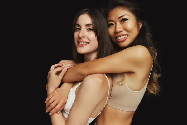 Portrait of young Asian woman hugs her friend from behind while they posing in underwear against black studio background. Concept of beauty and fashion, support, unity, wellness, body, diversity.