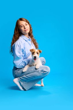 Çekici, uzun saçlı güzel bir kız yerde oturmuş, parlak mavi stüdyo geçmişine rağmen sevgili Jack Russel köpeğine sarılıyor. Hayvanlar ve sahipleri, dostluk, evcil hayvan bakımı kavramı. Ad