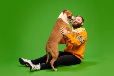 Turuncu tişörtlü adam, canlı yeşil stüdyo arka planına karşı arka ayakları üzerinde duran köpeğine mutlulukla sarılıyor. Hayvanlar ve sahipleri, dostluk, evcil hayvan bakımı kavramı. Ad