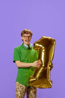 Genç kızıl saçlı bir adam, sıradan giyinmiş bir öğrenci, mor arka planda altın bir numaralı balonu tutuyor. Eğitim ve öğrenim kavramı, beceriler, bilgi, mezuniyet. Ad