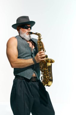 Klasik giyimli sakallı, olgun bir adam beyaz stüdyo arka planında altın saksafon çalıyor. Müzik anlayışı, festivaller, çağdaş sanat, hobi, yaratıcılık. Ad