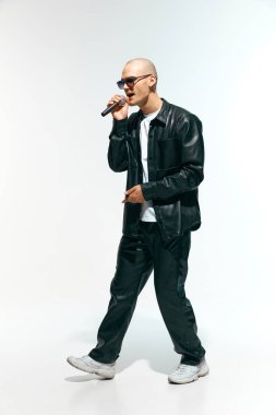 Koyu renk güneş gözlüklü, siyah deri ceketli, beyaz stüdyo arka planında mikrofon tutan kel bir adam. Müzik anlayışı, festivaller, çağdaş sanat, hobi, yaratıcılık. Ad