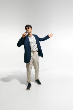 Genç adam gündelik kıyafetler giyip güneş gözlüğü takıyor mikrofon tutuyor ve şarkı söylüyor, beyaz stüdyo arka planına karşı gösteri yapıyor. Müzik anlayışı, festivaller, çağdaş sanat, hobi, yaratıcılık. Ad