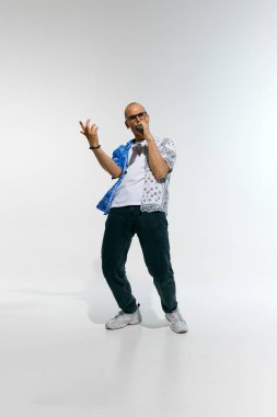 Gözlüklü, mavi ve beyaz desenli gömlekli genç adam, mikrofona enerji dolu bir şekilde beyaz stüdyo arka planında şarkı söylüyor. Müzik anlayışı, festivaller, çağdaş sanat, yaratıcılık.