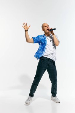 Moda kıyafeti giymiş sanatçı genç adam, vokalist, rapçi mikrofonla beyaz stüdyo arka planına karşı enerjik bir şekilde şarkı söylüyor. Müzik anlayışı, festivaller, çağdaş sanat, hobi, yaratıcılık. Ad