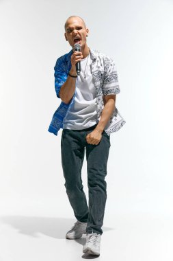 Artistik genç adam, moda kıyafeti giymiş, müzisyen, rapçi enerjik bir şekilde mikrofonla beyaz stüdyo arka planına karşı şarkı söylüyor. Müzik anlayışı, festivaller, çağdaş sanat, hobi, yaratıcılık. Ad